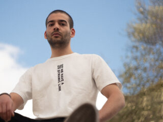 Eine Person, die ein weißes T-Shirt mit einem vertikalen QR-Code und dem Text „WIR HABEN EINEN DYNAMISCHEN LINK“ trägt, steht im Freien vor einem teilweise bewölkten blauen Himmel.