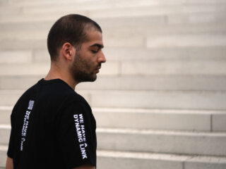 Eine Person mit kurzen Haaren steht seitlich auf einer Reihe heller Stufen und trägt ein schwarzes T-Shirt mit der Aufschrift „WIR HABEN EINE DYNAMISCHE VERBINDUNG“ in weißer Schrift auf der Rückseite.