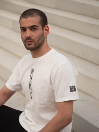 Ein Mann mit kurzen Haaren und Bart trägt ein weißes T-Shirt mit vertikalen schwarzen QR-Codes und Text und sitzt auf hellen Stufen.