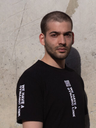Ein Mann mit rasiertem Kopf und Bart trägt ein schwarzes T-Shirt mit dem Text „WE HAVE A DYNAMIC LINK“ auf Ärmel und Brust und steht vor einer strukturierten Betonwand.
