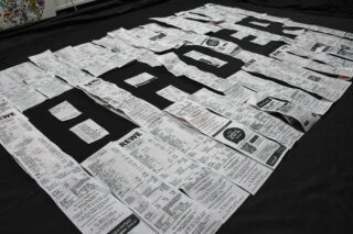 Ein großes schwarzes Tuch mit weißen Quittungen, die in dicken Blockbuchstaben ein Wort bilden; die Quittungen stammen aus verschiedenen Geschäften und überlappen sich teilweise.