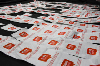 Auf einem dunklen Untergrund liegen zahlreiche weiße Papiertüten mit roten „REWE“-Logos ausgebreitet.