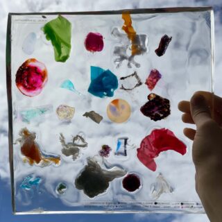 Eine Hand hält vor einem hellen Himmelshintergrund eine transparente quadratische Platte, in die verschiedene bunte, unregelmäßige Fragmente und Objekte eingebettet sind.