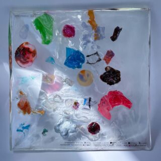 Ein durchsichtiger quadratischer Block mit verschiedenen bunten Plastikfragmenten und -objekten, angeordnet auf einer weißen Oberfläche mit sichtbaren Schatten an den Rändern.