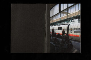 Passagiere warten auf Bänken auf einem Bahnsteig neben einem weißen Hochgeschwindigkeitszug in einem Bahnhof mit großen Fenstern und Deckenlicht.