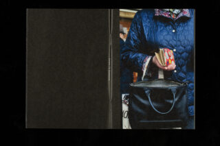 Eine Person in einer blauen Steppjacke hält eine gepunktete Brieftasche und eine schwarze Handtasche und steht vor einem überwiegend schwarzen Hintergrund.