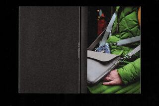 Eine Person in einem grünen Mantel sitzt mit einer grauen Handtasche und einem gemusterten Beutel auf dem Schoß, eine Hand ruht darauf, nur Oberkörper und Hand sind sichtbar.