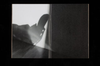 Der Schatten eines Stuhls und eines Teils einer Person wird auf eine weiße Wand und einen Holzboden geworfen, wobei die Szene durch die Mittelfalte eines offenen Buches oder einer Zeitschrift geteilt wird.