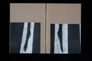 Ein hellbraunes Hardcoverbuch ist geöffnet und zeigt ein abstraktes Schwarzweißbild eines schattenhaften Beins und Fußes, das sich über beide Buchdeckel erstreckt.