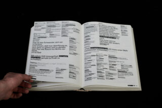 Eine Hand hält ein Buch aufgeschlagen, das Seiten mit deutschem Text zeigt, in denen Abschnitte hervorgehoben und fett gedruckt sind und die in einem blockartigen, überlappenden Stil formatiert sind.