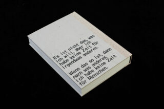 Ein geschlossenes Buch mit beigem Einband zeigt deutschen Text, der mit schwarzer Tinte auf schwarzem Hintergrund gedruckt ist.