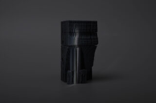 Ein dunkles, rechteckiges, blockartiges Objekt mit geschichteten, strukturierten Oberflächen steht aufrecht vor einem schlichten, dunklen Hintergrund.