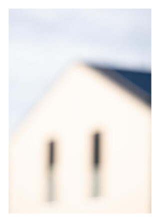 Ein verschwommenes Bild eines hellen Gebäudes mit einem dreieckigen Dach und zwei vertikalen Fenstern unter einem blassen Himmel.