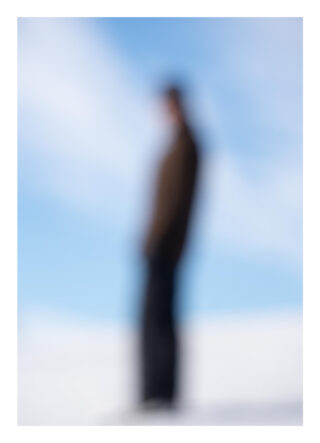 Ein verschwommenes Bild einer Person, die im Freien vor einem hellen, bewölkten Himmel steht.