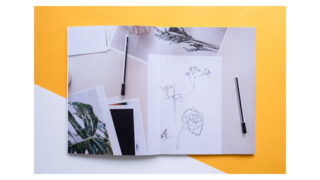 Ein offenes Skizzenbuch mit botanischen Strichzeichnungen und losen Fotos liegt auf einer gelb-weißen Oberfläche, daneben liegen zwei schwarze Stifte.