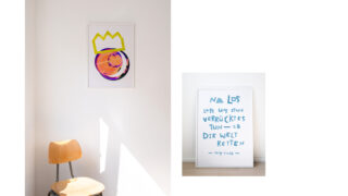 An einer Wand steht ein Holzstuhl mit zwei gerahmten Kunstwerken: eines mit abstrakten Formen und einer Krone darüber und ein anderes mit deutschem Text in Blau auf weißem Hintergrund.
