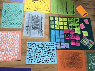 Auf einer Holzoberfläche sind verschiedene Blätter farbigen Papiers mit abstrakten schwarz-weißen Mustern und Pinselstrichen angeordnet.