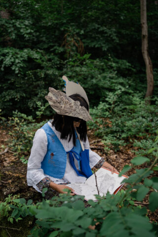 Eine Person mit einem großen, dekorativen Papierhut sitzt draußen in einem Waldstück und schaut auf ein offenes Skizzenbuch auf ihrem Schoß.