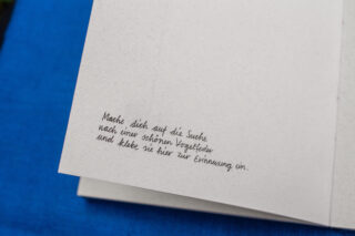 Auf einer handgeschriebenen deutschen Notiz in einem Notizbuch steht auf einer leeren Seite: „Mache dich auf die Suche nach einer schönen Vogelfeder und klebe sie hier zur Erinnerung ein“.