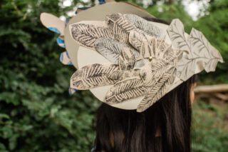 Eine Person mit langem, dunklem Haar trägt vor einem unscharfen Außenhintergrund eine große Kopfbedeckung aus mehrschichtigen Papierausschnitten mit Blattmuster.