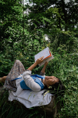 Eine Person liegt auf einer Bank im Freien auf einer Rasenfläche, lächelt und liest ein Buch oder Notizbuch, umgeben von grünem Laub.