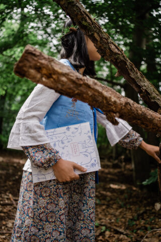 Eine Person in gemusterter Kleidung hält einen dekorativen Ordner und steht zwischen Bäumen, teilweise verdeckt durch einen Ast in einem Waldstück.