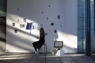 Eine Person sitzt auf einem Hocker vor einer Kunstinstallation an einer weißen Wand mit Stücken aus verschiedenen Medien, einem Bildschirm auf einem Podest und Sonnenlicht, das Schatten über die Szene wirft.