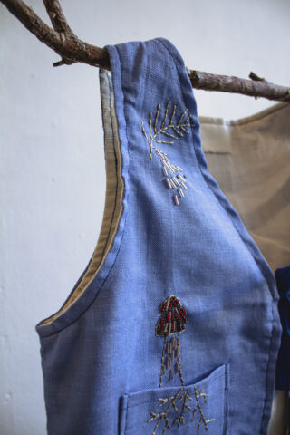 Eine blaue Leinenschürze mit gestickten Mustern, darunter ein roter und brauner Pilz und kleine Zweige, hängt an einem natürlichen Holzast.