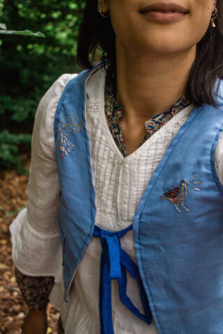 Person, die eine weiße Bluse mit einer blauen bestickten Weste und einem gemusterten Kragen trägt und im Freien mit viel Grün im Hintergrund steht.