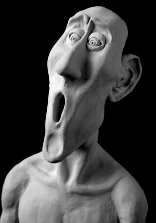 Eine monochrome Skulptur einer menschenähnlichen Figur mit übertriebenen Gesichtszügen, großen Augen und offenem Mund, die Überraschung oder Schock auszudrücken scheint.