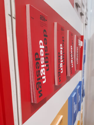 Mehrere rote Bücher mit dem Wort „Design“ in fetten schwarzen Buchstaben stehen vertikal auf einem rot-weißen Regal. Darunter sind blaue und gelbe Bücher zu sehen.