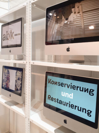 Vier Apple-Desktop-Monitore stehen auf weißen Regalen. Zwei Bildschirme zeigen deutschen Text: „abk“ und „Konservierung und Restaurierung“. Die anderen beiden zeigen abstrakte Bilder.
