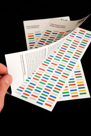 Eine Hand blättert durch bedruckte Blätter mit mehrfarbigen rechteckigen Mustern und Textblöcken, alle vor einem schwarzen Hintergrund.