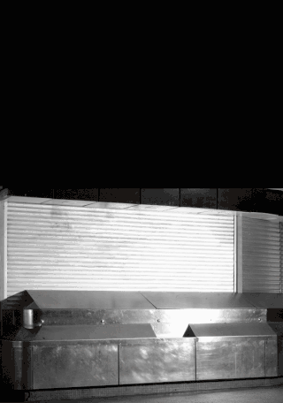 In einem dunklen, unbeleuchteten Raum steht eine große, leere Edelstahl-Ausstellungstheke vor einem geschlossenen weißen Rolltor.