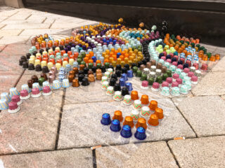 Reihen bunter Kaffeepads sind auf Steinfliesen im Außenbereich angeordnet und bilden ein dekoratives, wirbelndes Muster.