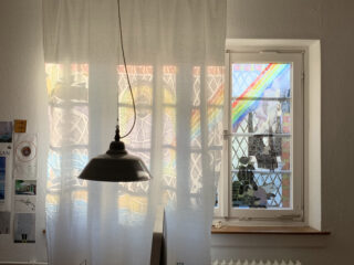 Vor einem Fenster mit bunten Buntglasfenstern hängt ein weißer Vorhang, im Vordergrund schwebt eine schwarze Pendelleuchte.