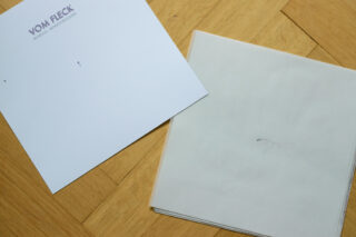 Ein einzelnes Blatt Papier mit der Aufschrift „Vom Fleck“ liegt neben einem Stapel leerer weißer Blätter auf einem Holzboden.