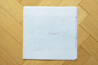 Auf einem hellen Holzparkettboden liegt ein Stapel durchscheinender Papiere mit schwachen Bleistift- und rosafarbenen Markierungslinien.