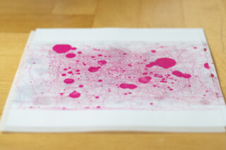 Ein Blatt Papier auf einer Holzoberfläche mit rosa Tintenspritzern und Linien, die ein abstraktes Muster bilden.