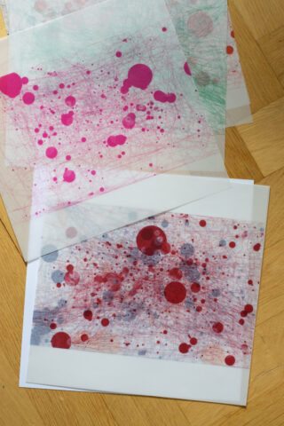 Mehrere durchscheinende Blätter und ein Papier zeigen abstrakte Muster aus Linien und roten Kreisen, die auf einem Holzboden ausgelegt sind.