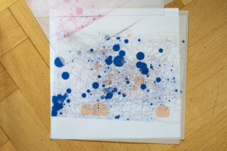 Auf einem Holzboden liegt ein Blatt Transparentpapier mit blauen und pfirsichfarbenen Tintenflecken und Kritzeleien, teilweise bedeckt von einem weiteren durchscheinenden Blatt mit rosa Flecken.