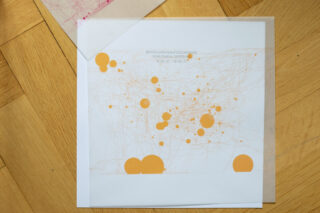 Ein weißes Blatt Papier mit schwachen orangefarbenen Linien und verschiedenen orangefarbenen Kreisen unterschiedlicher Größe liegt auf einem Holzboden und ist teilweise mit einer transparenten Folie bedeckt. Oben ist deutscher Text aufgedruckt.