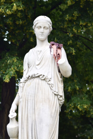 Vor grünen Bäumen steht die Marmorstatue einer Frau mit einem Krug in der Hand und einem Stück rosa Stoff über dem erhobenen Arm.