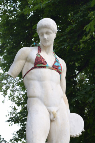 Eine klassische Marmorstatue einer nackten männlichen Figur in einem bunten Bikinioberteil mit Bäumen und Grün im Hintergrund.