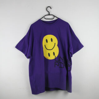 Ein lila T-Shirt mit zwei großen gelben Smileys und schwarzem Text auf der Vorderseite hängt auf einem schwarzen Kleiderbügel vor einer weißen Wand.