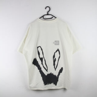 An einer Wand hängt ein weißes Kurzarm-T-Shirt mit einer großen abstrakten schwarzen Grafik und einem kleinen gemusterten Quadrat auf der Rückseite.