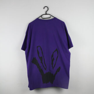 Ein lila T-Shirt hängt auf einem schwarzen Kleiderbügel vor einer schlichten weißen Wand. Auf der Rückseite ist eine große schwarze abstrakte Grafik abgebildet, die einem Gesicht oder Symbol ähnelt.