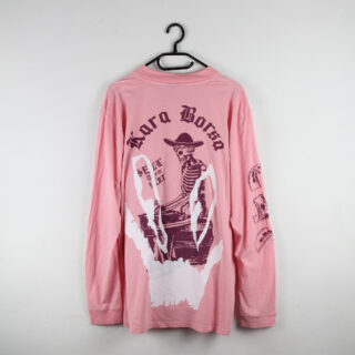 Ein rosa Langarmshirt mit einer großen Grafik eines Cowboy-Skeletts, dem Text „Kora Borra“ und weiteren Motiven auf den Ärmeln, das auf einem schwarzen Kleiderbügel vor einer weißen Wand hängt.