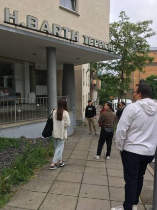 Vier Personen stehen vor einem Gebäude mit dem Schild „H. BARTH TEPPICHE“ über dem Eingang. Eine Person blickt zum Gebäude, die anderen stehen in der Nähe auf dem Bürgersteig.