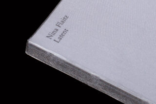Eine Nahaufnahme eines Buches mit weißem Einband und dem Titel „Nina Flaiz Latent“, fotografiert vor einem schwarzen Hintergrund.